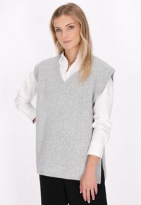 Světlé šedé bezrukávové svetr s výst�řihem do V a bočními rozparky, nošený přes bílou košili. Měkká textura, minimalistický design, uvolněný střih.