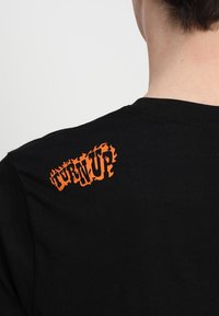 Černé bavlněné tričko s texturou grafiky ve stylu oranžového plamene "TURN UP" potisknuté na horní části zad, kulatým výstřihem a krátkými rukávy.