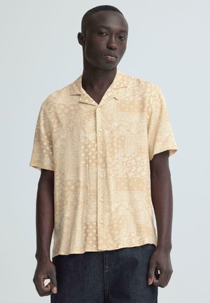 Mężczyzna w beżowej koszuli na guziki z krótkim rękawem z wzorem oraz ciemnych jeansach, stojący na tle jednolitego, jasnoszarego tła.