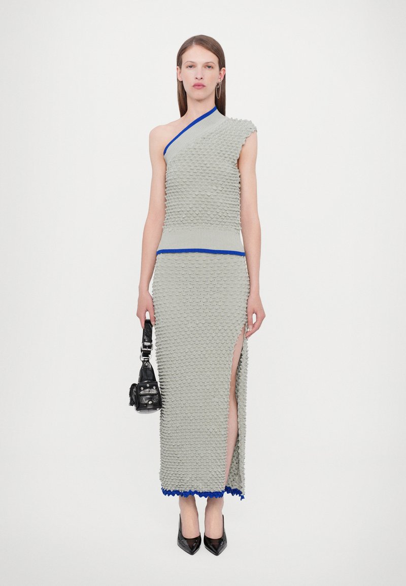 Vestido de punto gris con patrón de escamas texturizado, hombro asimétrico, acentos azules y abertura alta en el muslo, combinado con un pequeño bolso negro.