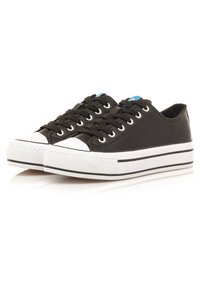 Zapatillas de lona negras con suelas y capuchones de goma blancos. Presentan cordones planos negros y un acento azul cerca de la lengüeta. Textura sutil.
