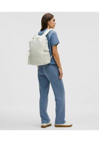 Mochila blanca con cierre de cremallera, correas ajustables y bolsillo delantero. El modelo lleva puesta una camisa azul y pantalones, con zapatillas blancas.