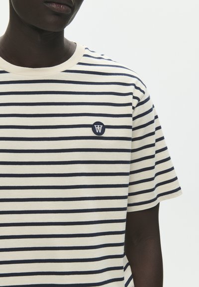 Homme portant un t-shirt à manches courtes rayé horizontalement crème et bleu marine avec un petit logo circulaire sur la poitrine.