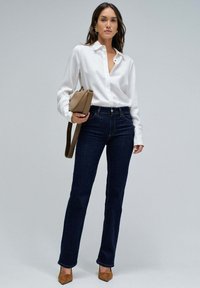 Mujer vestida con camisa blanca de botones, jeans azul oscuro, tacones marrones y sosteniendo un bolso beige con pendientes dorados frente a un fondo liso.