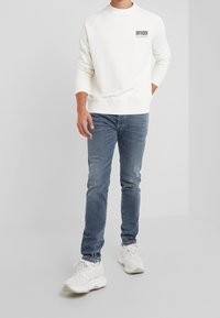 Sudadera blanca con el logo "DRYKORN", combinada con jeans ajustados azul y zapatillas de color claro. Tejido suave, corte relajado, diseño de cuello redondo.