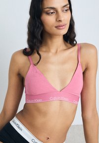 Roza braletka iz mehkega materiala s trikotno obliko, nastavljivimi naramnicami in pasom z logotipom Calvin Klein. Tekstura je gladka z rahlimi vzorci.