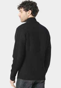 Černý pletený svetr s ribbed texturou, vysokým límcem a výraznými horizontálními a vertikálními vzory na zádech a rukávech.