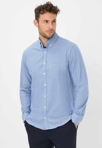 Man in een lichtblauw, klein geruit overhemd met lange mouwen, staand met zijn handen in de zakken van een donkere broek, kijkend opzij.