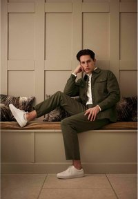 Olijfgroen jack en bijpassende broek van textuurstof, gecombineerd met een wit shirt en witte sneakers, zittend op een houten bank.
