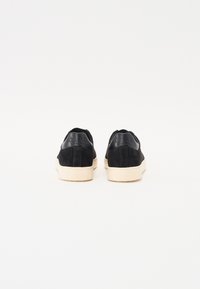 Sneakers in suede nera con linguetta nera liscia, suola in gomma crema e dettaglio del logo embossato sul tallone. Design semplice e minimalista.