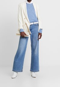 Vit blazer över blått texturerat tröja, högmidjade blå vida jeans, vit bälte och vita anklar stövlar. Håller en liten clutch.