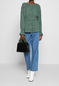 Blusa verde con estampado floral blanco, escote con fruncido y mangas largas acampanadas. Combinada con jeans de pierna recta azul claro y un bolso negro texturizado.