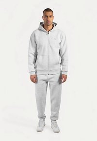Felpa grigia chiaro con zip e tasca a marsupio, abbinata a pantaloni della tuta coordinati, entrambi realizzati in tessuto morbido. Presenta una cappuccio con coulisse.