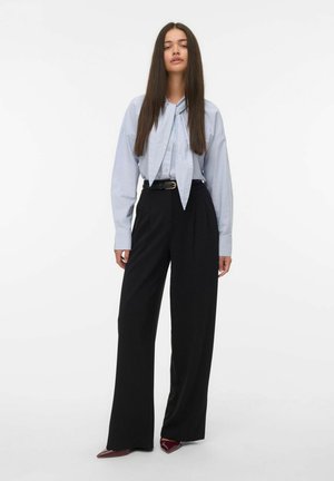 Jonge vrouw met lang steil haar, gekleed in een lichtblauwe blouse met een stropdas, hoge taille zwarte wijde broek en donkere puntige schoenen.