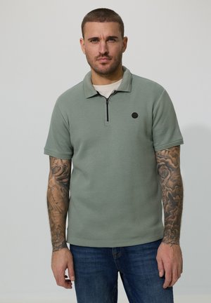 Mężczyzna z wytatuowanymi ramionami, ubrany w miętowozieloną, krótkorękawową koszulkę polo na zamek i niebieskie dżinsy, stojący na tle jednolitego jasnego tła.