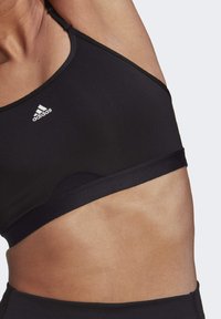 Črn športni nedrček z nastavljivimi naramnicami, ki ima zaobljen izrez in bel logo "adidas" na zgornji levi strani. Gladek, raztegljiv material.