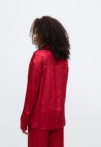 Cămașă lungă cu mâneci lungi din satin roșu, cu un model geometric, guler ascuțit și manșete cu nasturi, văzută din spate. Textură netedă și lucioasă.