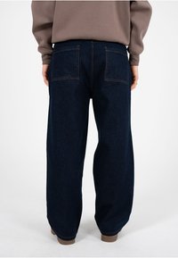 Pantalones de mezclilla azul oscuro de pierna ancha con dos bolsillos traseros y detalles de costura visibles. Llevados con una sudadera marrón claro.