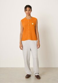 Oranje mouwloze gebreide cardigan met een ronde hals, knopen aan de voorkant en een klein label. Gecombineerd met grijze ruimzittende sweatpants en bruine schoenen.