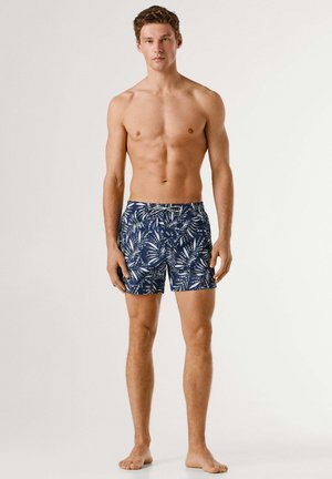 Męski model stojący boso, ubrany w granatowe kąpielówki z białym tropikalnym wzorem liści na jednolitym tle.