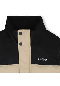 Veste matelassée noire et beige, avec un col montant, des boutons-pression et le logo "HUGO" en blanc. Tissu doux et texturé.