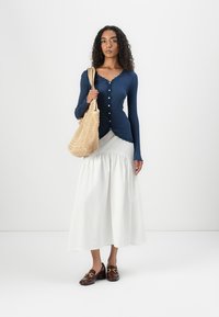 Haut à manches longues en maille côtelée bleue marine, jupe blanche à volants, sac tissé beige et chaussures marron foncé avec des accents métalliques.