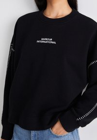 Černá mikina s bílým textem "Barbour International", límeček Crew neck, volný střih a bílé švy podél švů rukávů.