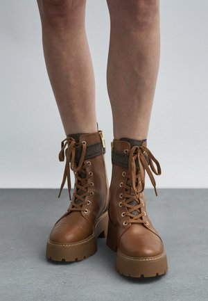 Bottines en cuir marron avec un talon épais, devant à lacets, revers en tissu gris et accents en métal doré. Surface texturée remarquable.
