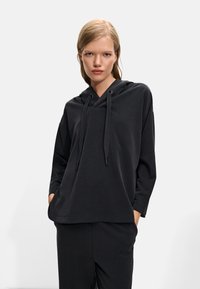 Hanorac negru cu glugă, cu o croială lejeră, mâneci lungi și șnur la gât. Textură moale și design minim, asortat cu pantaloni corespunzători.