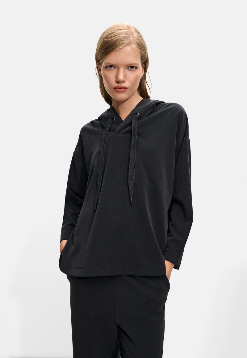 Hanorac negru cu glugă, cu o croială lejeră, mâneci lungi și șnur la gât. Textură moale și design minim, asortat cu pantaloni corespunzători.