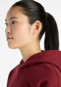 Burgundy hoodie with a large front pocket and drawstring hood, worn by a person with straight black hair tied back, viewed from the side.