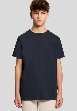 Tienerjongen met kort bruin haar, gekleed in een effen zwart T-shirt met ronde hals en beige broek, staand tegen een grijze achtergrond.