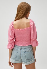 Lyserød smocket bluse med pufærmer, firkantet halsudskæring og flagrende kanter. Sammen med lyse jeansshorts med omdrejede manchetter.