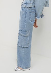 Persona che indossa jeans in denim azzurro chiaro decorati con una grande tasca laterale, abbinati a una giacca di jeans coordinata e scarpe piatte argentate.