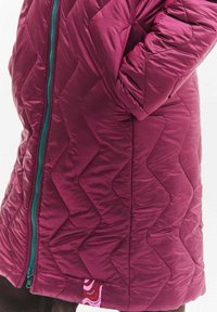 Bordó prošívaný péřový kabát s přiléhavým střihem, vybavený tyrkysovým zipem a růžovou značkovou visačkou na lemu. Výrazný vzor ve tvaru šipky.