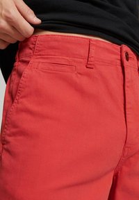 Pantalones cortos de algodón rojos con frente plano y bolsillo lateral, con un cierre de botón estándar en la cintura y un diseño de corte recto.