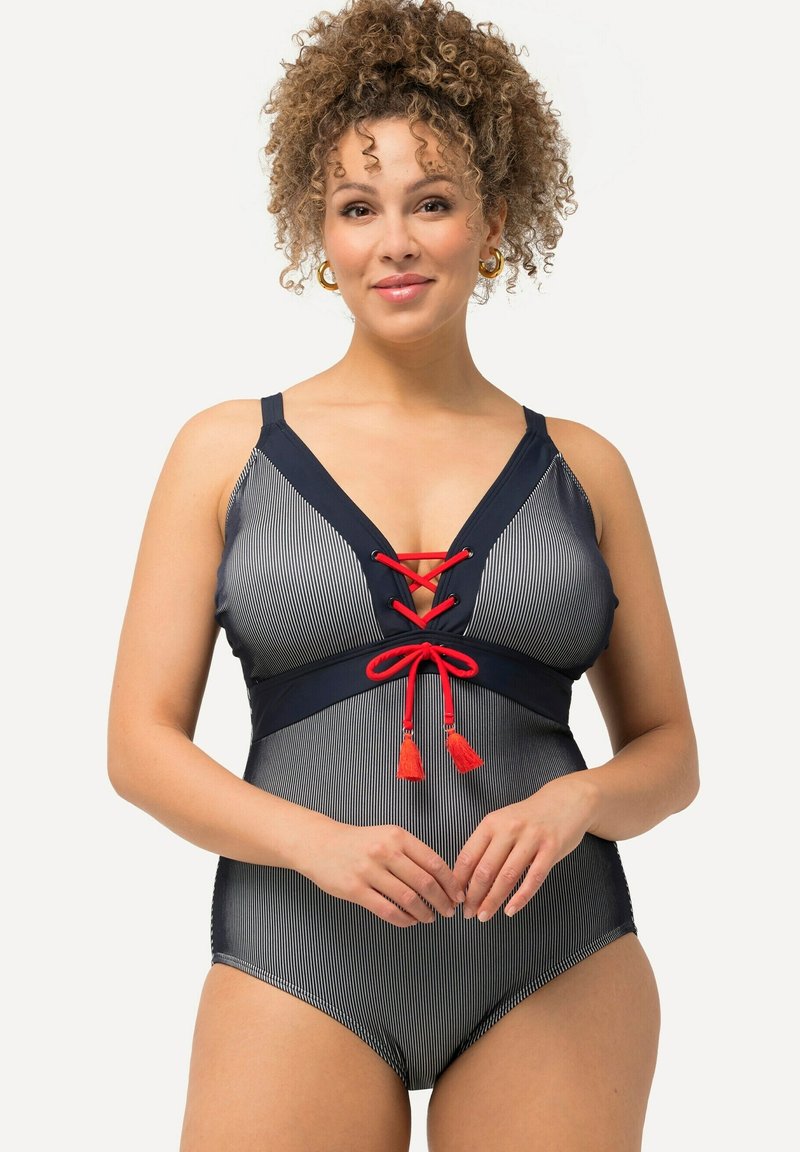 Costume da bagno intero a righe blu navy e bianche con scollatura a V profonda, dettagli in pizzo rosso con lacci e accentuazioni in contrasto con pompon. Spalline regolabili.