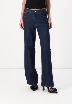 Mujer con vaqueros de pierna ancha de rayas finas azul oscuro combinados con tacones altos negros de punta puntiaguda con detalles de lazo.