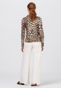 Donna con capelli rossi raccolti in una coda di cavallo, che indossa una maglietta a maniche lunghe con stampa leopardata, pantaloni bianchi a gamba larga e scarpe nere aperte sul retro, in piedi con la schiena rivolta.