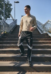 Camisa de manga larga beige con logo, pantalones deportivos negros con detalles en blanco y rojo, zapatos negros, bajando escaleras bajo un cielo azul claro.