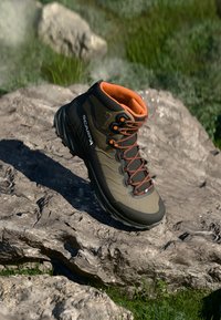 Buty do wędrówek z pomarańczowym wykończeniem i sznurówkami na skalistym terenie, z nazwą marki "Scarpa" po boku, otoczone trawą i światłem słonecznym.