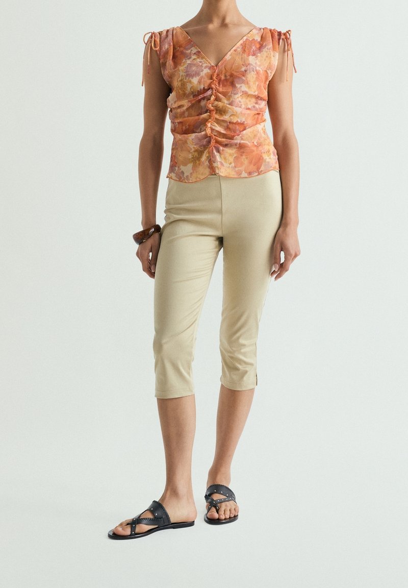 Femme portant un chemisier froncé à imprimé floral orange, un pantalon beige court, des sandales noires à clous et un bracelet marron, debout sur fond blanc.
