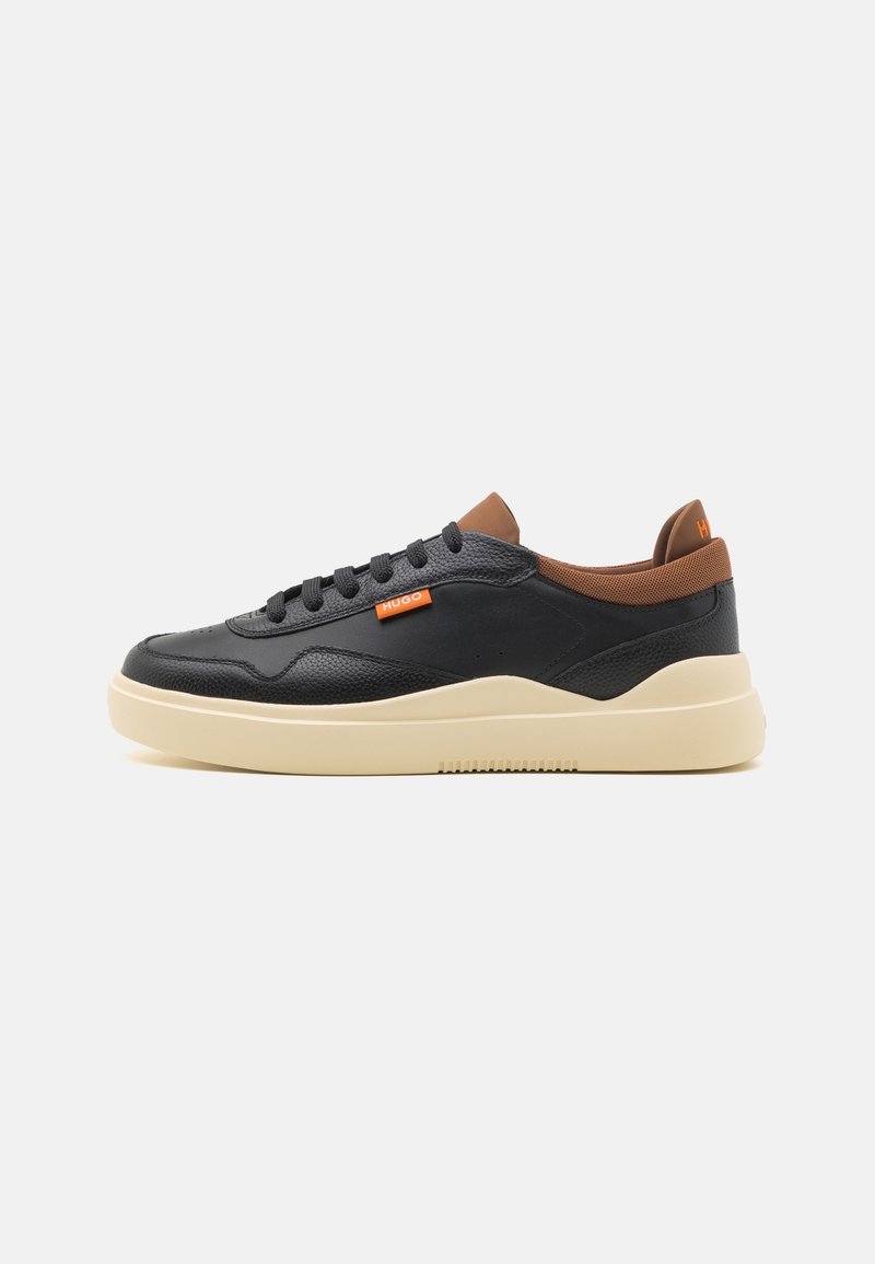 Baskets basses en cuir noir avec col marron, semelle beige, lacets noirs et étiquette de marque orange sur le côté et la languette du talon.