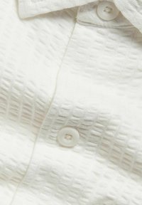 Witte textuurshirt met een subtiele ruitpatroon, voorzien van een klassieke kraag en een ronde witte knoopsluiting.