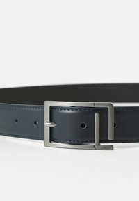 Cintura in pelle blu scuro con fibbia rettangolare argentata, texture liscia e fori equidistanti su uno sfondo bianco.