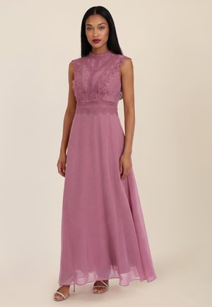Woman wearing a sleeveless mauve lace top evening gown with a flowing skirt and silver open-toe heels, standing against a plain background.
