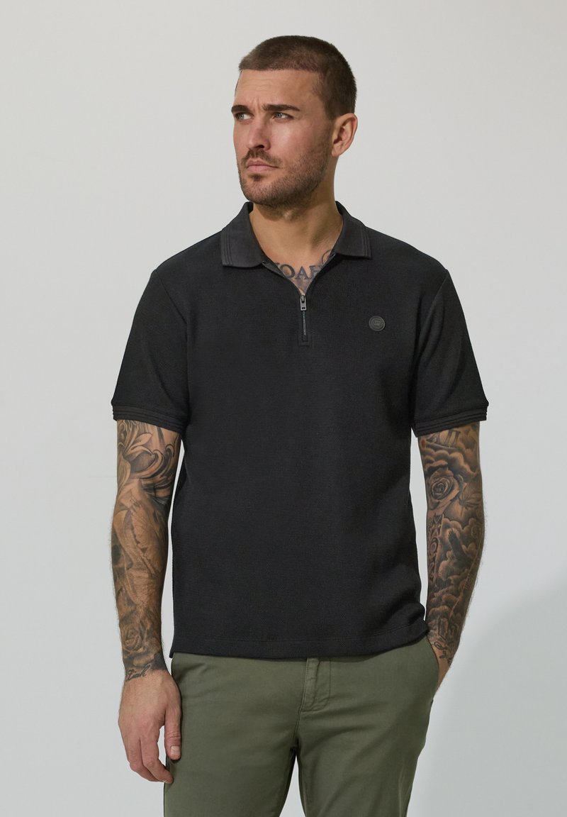 Homme aux cheveux courts et tatouages sur les bras, portant un polo noir à col zippé et un pantalon vert olive, regardant vers la gauche avec une main dans la poche.