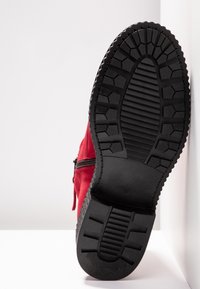 Semelle en caoutchouc noir avec des motifs de crampons sur une botte en daim rouge, présentée debout verticalement sur un fond blanc.