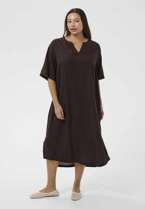 Kobieta w luźnej, ciemnobrązowej sukience do kolan z krótkimi rękawami oraz beżowych wsuwanych butach z plecionki, stojąca na jednolitym tle.