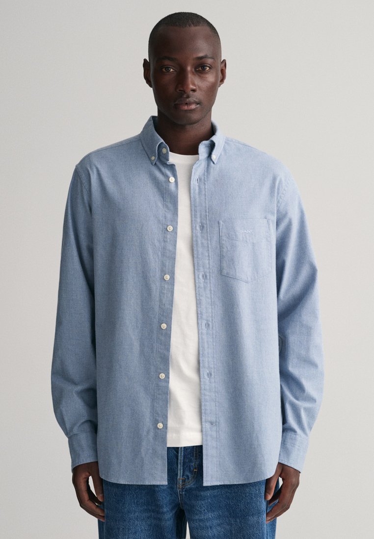Hombre con camisa de botones azul clara sobre una camiseta blanca y jeans azules, de pie contra un fondo liso claro.